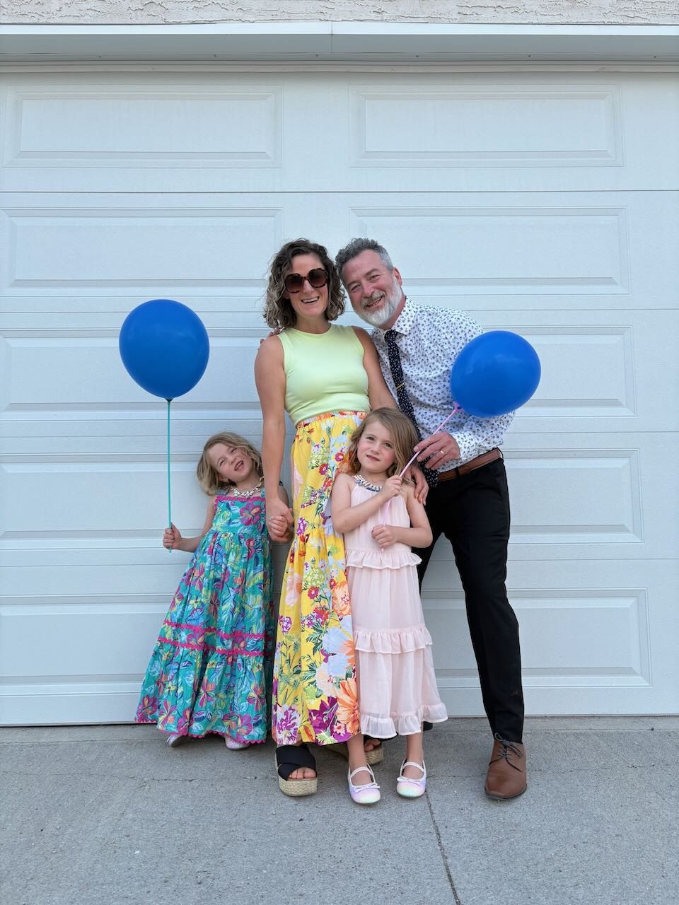 Matt and Megan Sodomsky with their two daughters, holding blue balloons in front of a white garage door